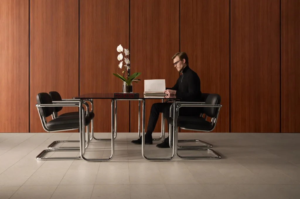 Modern professional working at a glass table surrounded by reissued SOM79 chairs in a minimalist interior with warm wood paneling and white orchids, echoing Halston’s iconic Olympic Tower aesthetic