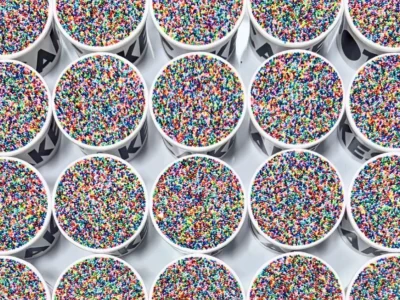 Overhead view of dozens of Dot Cakes arranged in neat rows, each served in a branded white cup and topped with a dense layer of colorful rainbow nonpareil sprinkles. The vibrant sprinkle-covered surfaces create a striking mosaic effect, highlighting the viral dessert’s playful, nostalgic aesthetic and signature crunchy topping