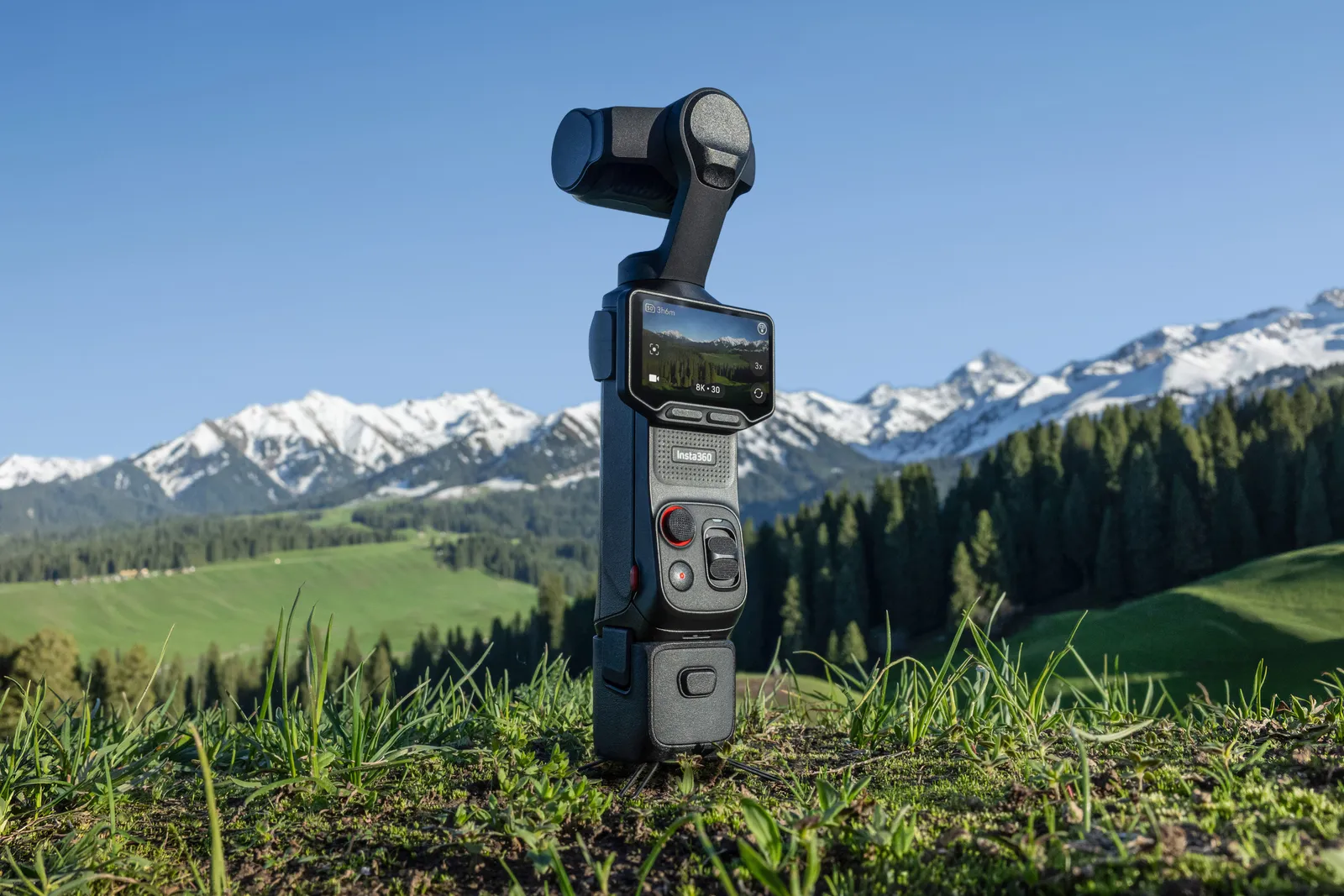 Insta360 handheld camera positioned outdoors on grassy terrain with snow-capped mountains in the background, illustrating its portability and suitability for travel, adventure, and landscape video