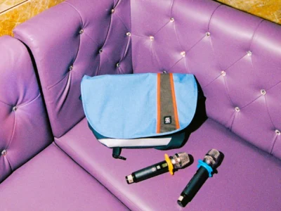 Blue Crumpler messenger bag resting on a tufted purple booth seat beside two handheld microphones in a karaoke lounge, highlighting the bag's contrasting orange accent stripe and urban lifestyle set