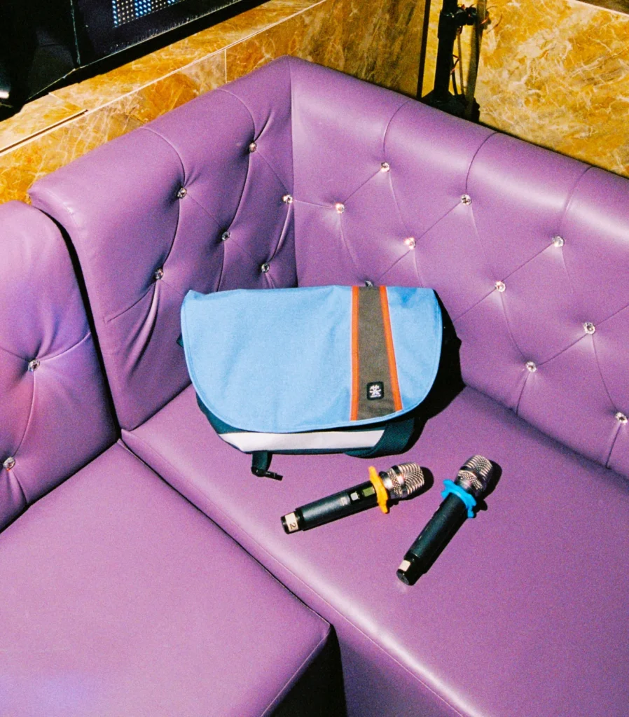 Blue Crumpler messenger bag resting on a tufted purple booth seat beside two handheld microphones in a karaoke lounge, highlighting the bag's contrasting orange accent stripe and urban lifestyle set