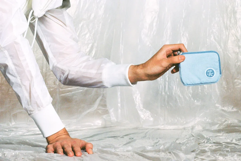 Hand holding a light blue Crumpler zip pouch against a translucent plastic backdrop, showcasing the compact organizer with a tonal embroidered logo and minimalist design