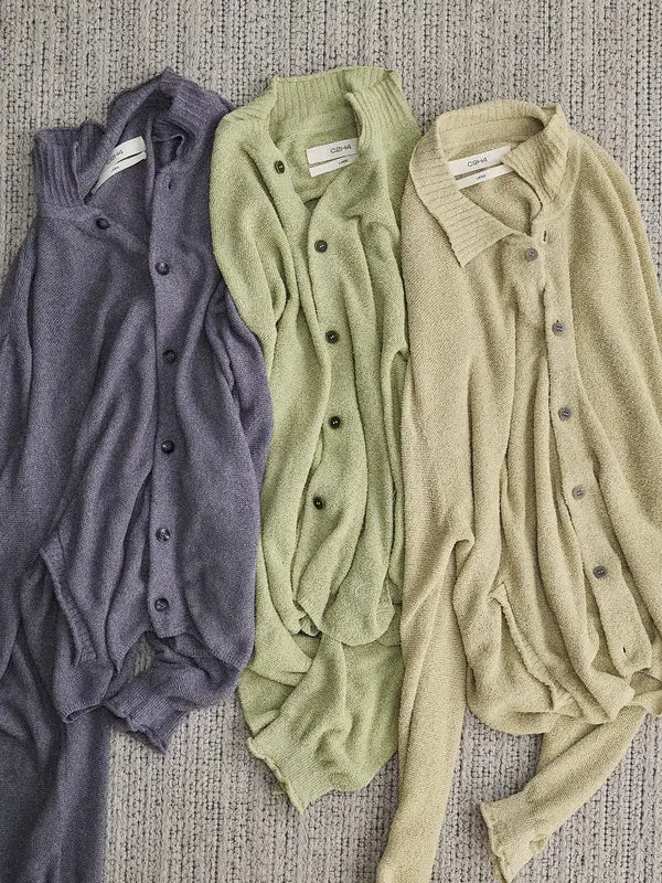 Three lightweight knit cardigans in charcoal, sage green, and beige laid side by side on a textured carpet, each featuring a button front, relaxed collar, and soft open-knit construction.