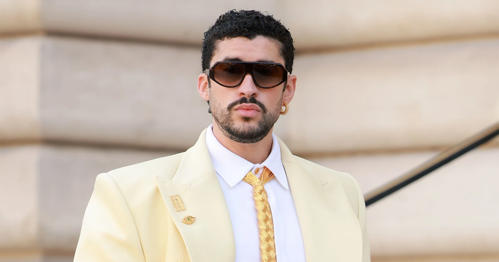 Close-up portrait of a man wearing oversized brown-tinted Gentle Monster sunglasses, styled with a cream tailored blazer, white shirt, braided gold tie, and gold jewelry against a softly blurred architectural backdrop.