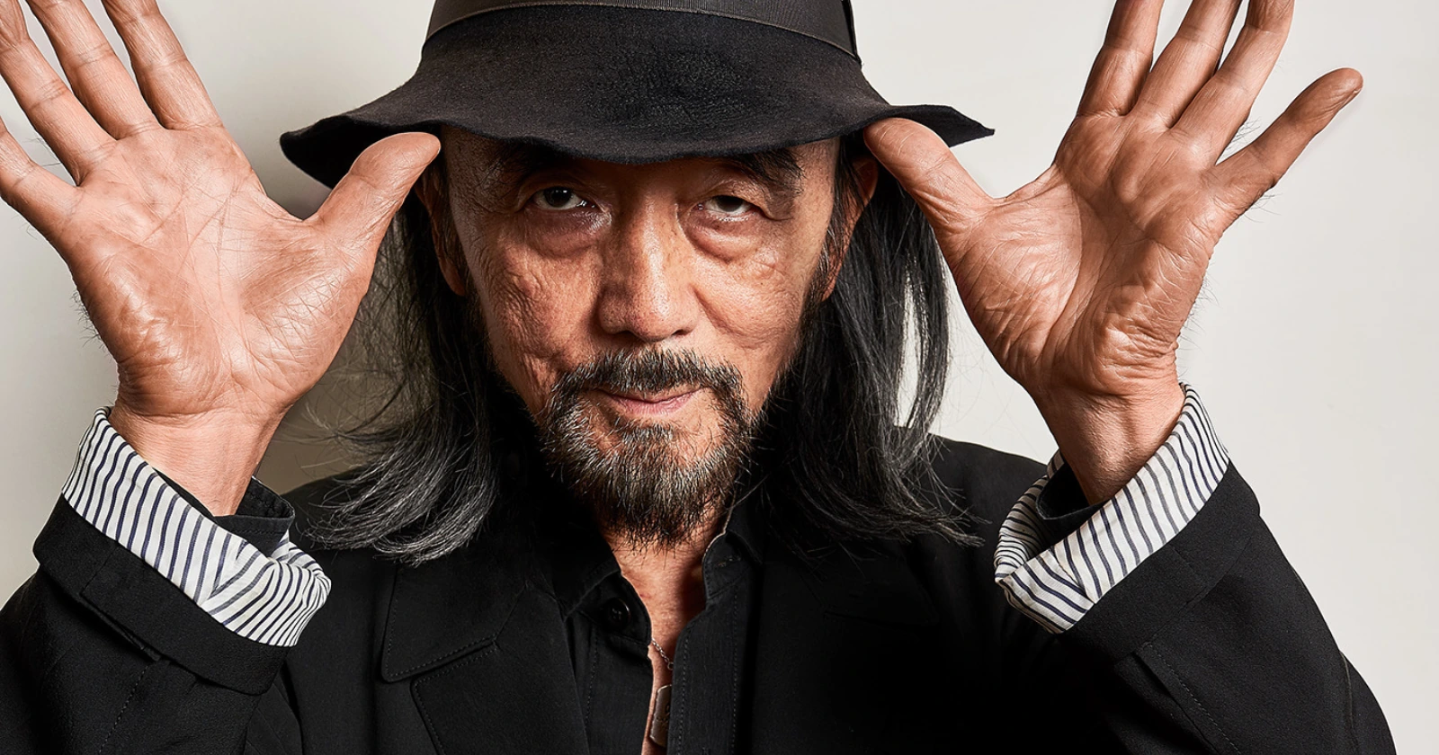 Yohji Yamamoto poses with both hands raised beside his face, wearing a black fedora, black jacket, and striped shirt cuffs. The close portrait highlights his expressive gesture, long gray hair, and signature monochromatic style against a plain background.