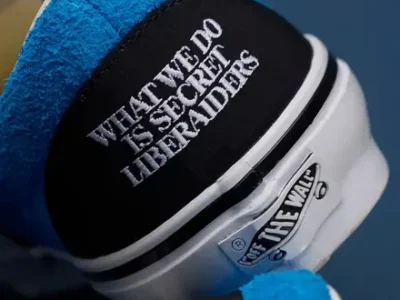 Close-up of the Vans heel showing white embroidered text reading “WHAT WE DO IS SECRET LIBERAIDERS” on a black suede panel, with bright blue suede collar lining and the classic Off The Wall heel tab