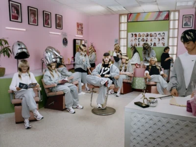 Modern hair salon with pastel pink walls where multiple people in matching gray Nike fleece sets relax in styling chairs, read magazines, chat with stylists, and wait beneath vintage hooded hair dryer