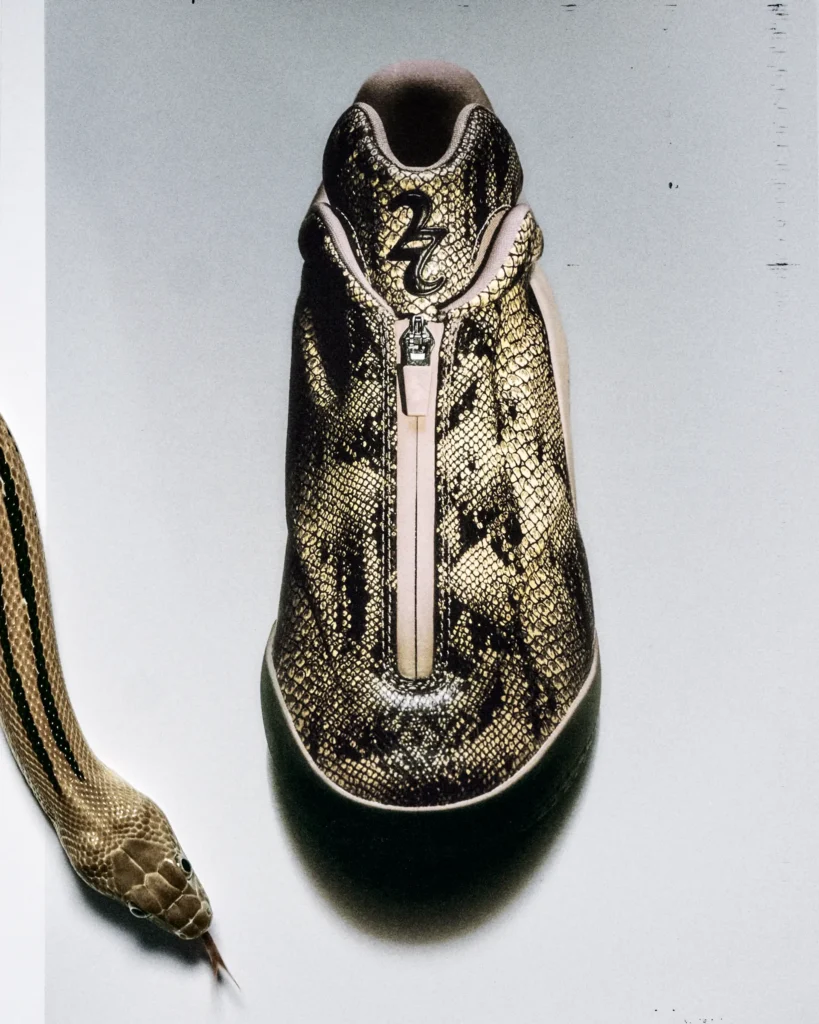 Front-facing view of the Converse SHAI 001 basketball shoe in a tan and brown snakeskin print. A full-length center zipper replaces traditional laces, while the padded collar features Shai Gilgeous-Alexander's "2" logo. A snake enters the frame from the lower left against a clean light-gray studio backdrop