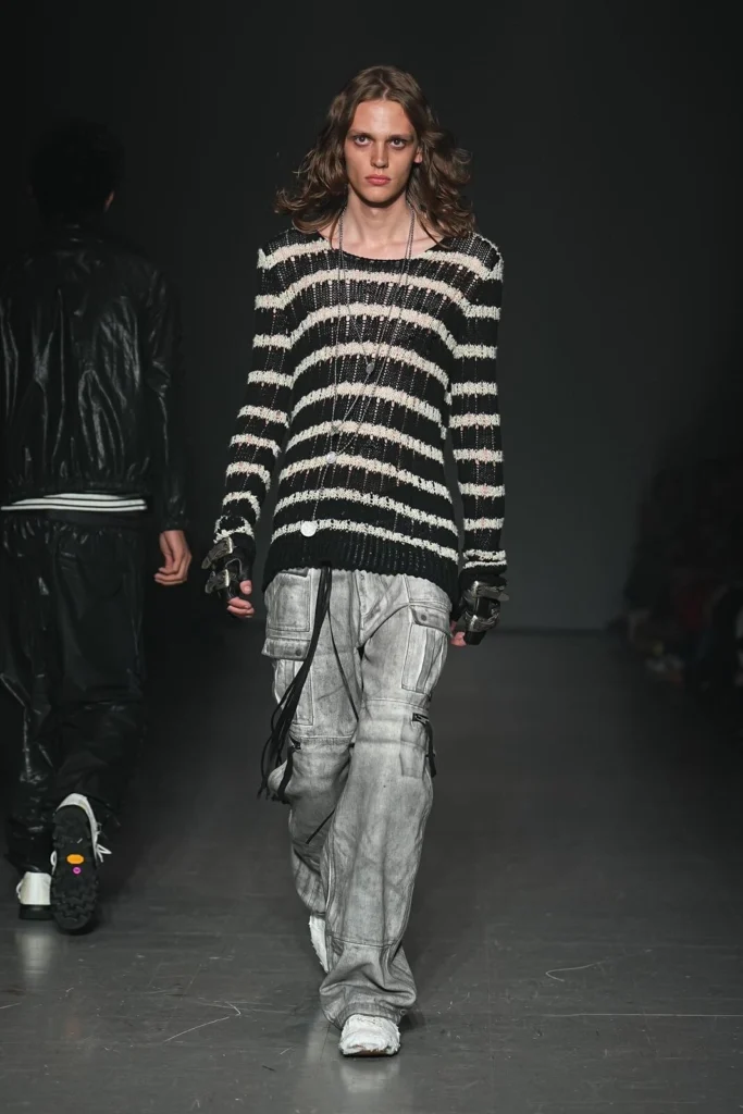 Model wearing a black-and-white striped knit sweater with oversized faded gray cargo pants, layered necklaces, fingerless gloves, and white sneaker on the runway
