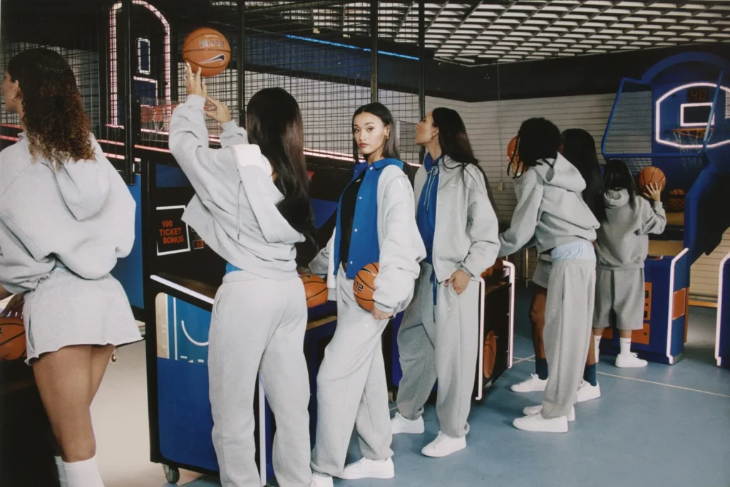 Group wearing coordinated gray Nike fleece and blue layers gathered around arcade basketball games, with one person holding a basketball while others take turns shooting in a neon-lit entertainment space