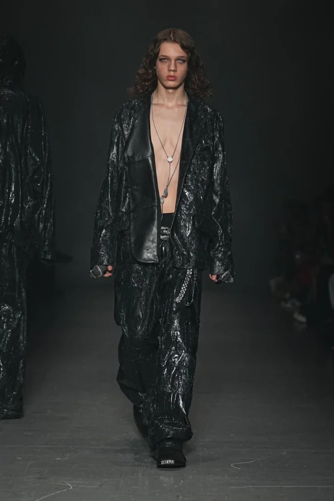 Model in a glossy black textured jacket and matching wide-leg pants worn open over a bare chest with layered pendant necklaces and black footwear