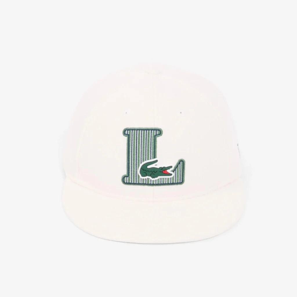White snapback cap featuring a green striped varsity-style "L" logo with the signature Lacoste crocodile embroidered across the base, shown against a clean white background