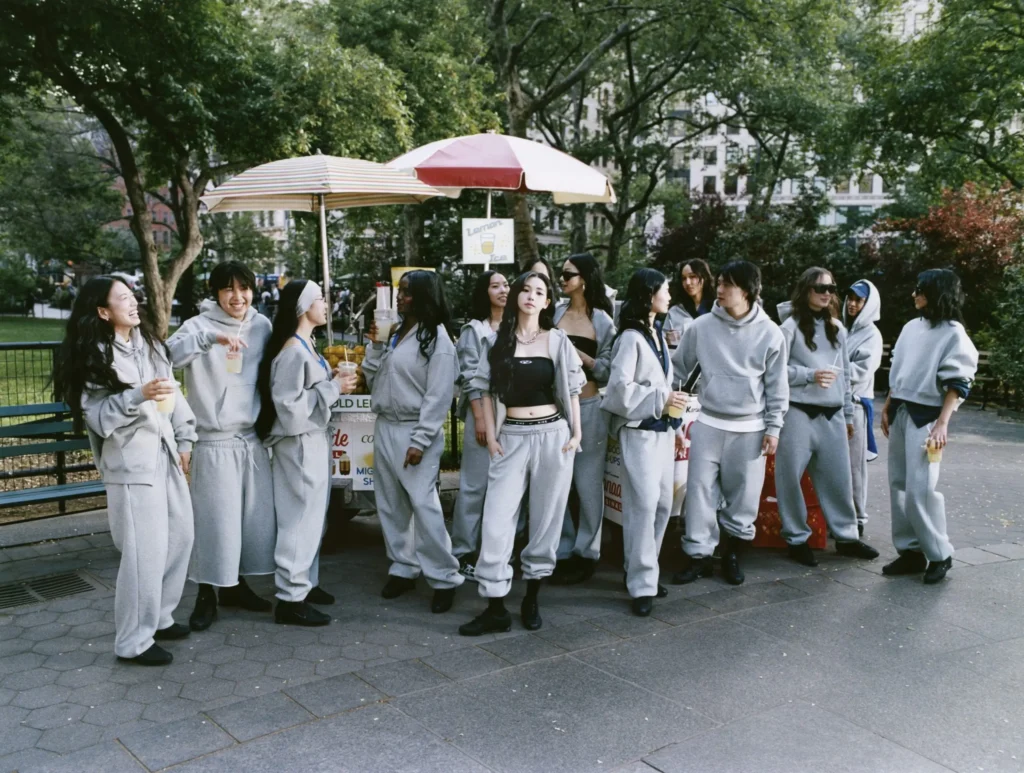 Large group dressed in matching gray Nike fleece outfits gathered around a city park lemonade cart, socializing and holding drinks while one person stands confidently in the center wearing a black crop top