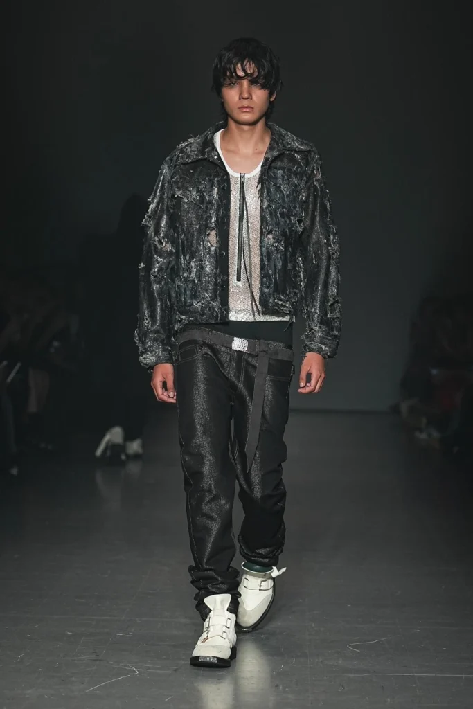 Model wearing a distressed charcoal cropped jacket over a white mesh top, slim black trousers with a hanging belt strap, and white buckle shoe