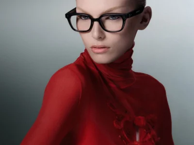Studio portrait of a model wearing black square smart glasses with clear lenses, styled in a fitted red high-neck top with textured detailing and slicked-back blonde hair against a muted gray background