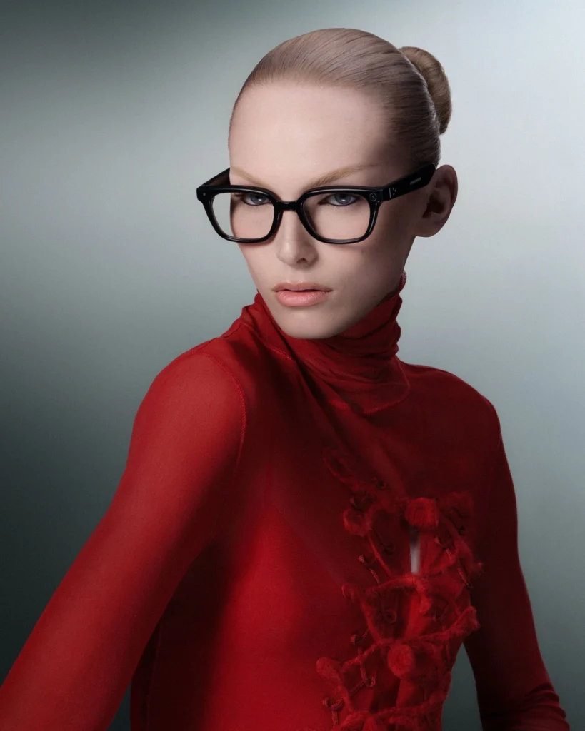 Studio portrait of a model wearing black square smart glasses with clear lenses, styled in a fitted red high-neck top with textured detailing and slicked-back blonde hair against a muted gray background