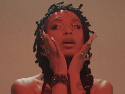 Close-up portrait of a person under warm red lighting with shoulder-length twisted hair, hands framing their face, glossy lips slightly parted, long embellished nails, stacked bracelets, multiple rings, and visible tattoos against a softly textured neutral background
