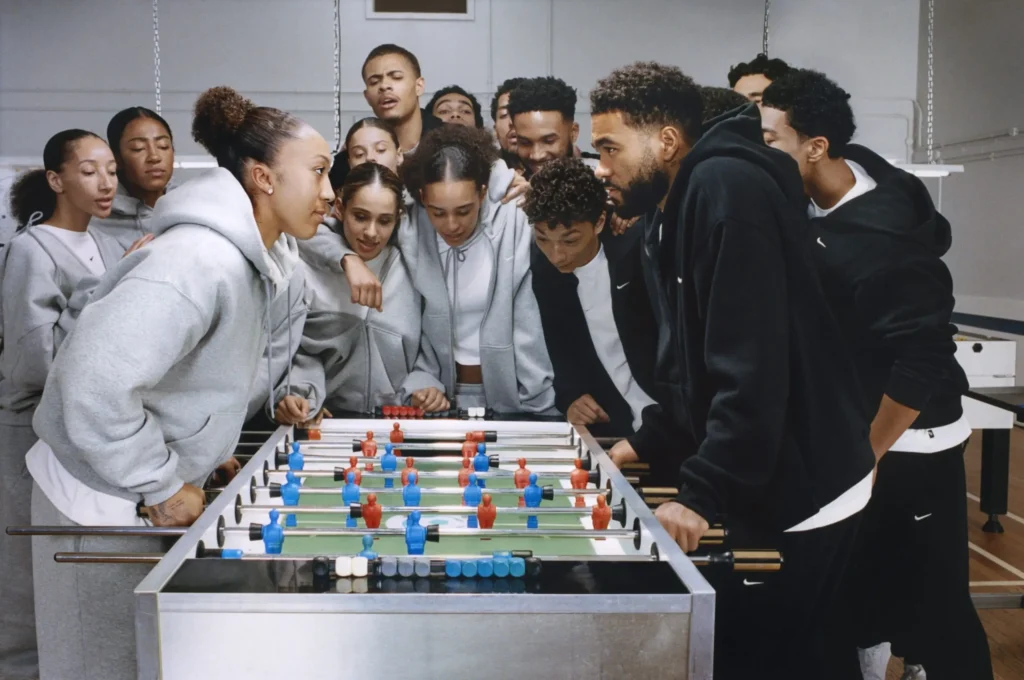 Group of athletes in gray and black Nike fleece hoodies gathered around a foosball table, leaning in and watching an intense match with focused expressions inside a recreation room
