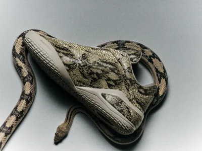 Side profile of the Converse SHAI 001 wrapped by a coiled snake. The shoe features an all-over tan snakeskin pattern with dark markings, a sculpted midsole, and zippered upper, photographed on a minimalist gray background