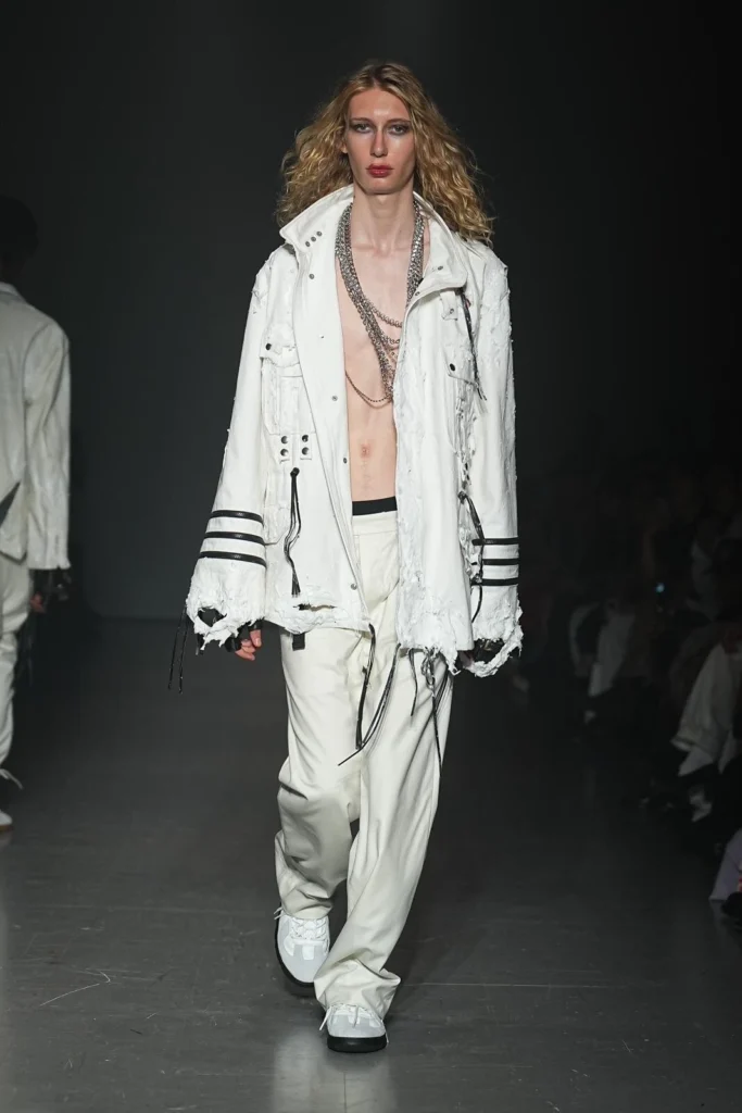 Model in an oversized distressed white jacket with matching relaxed trousers, layered silver chains, exposed chest, and white sneaker