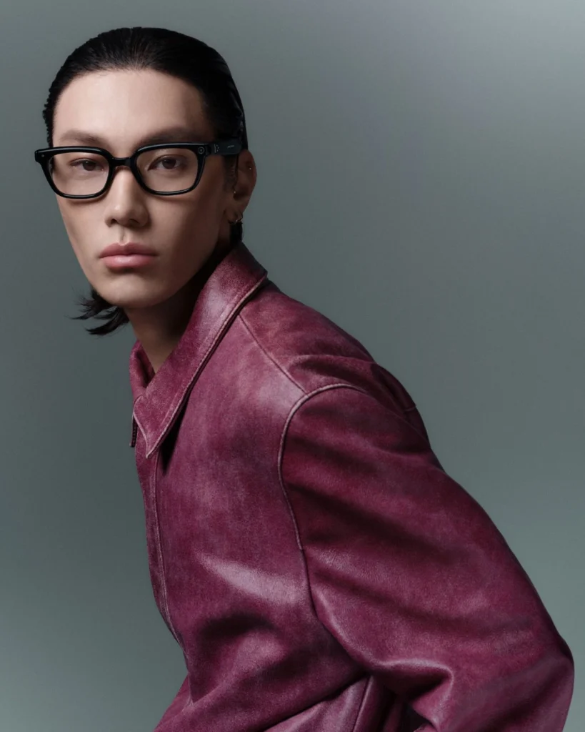 Studio portrait of a model wearing black rectangular smart glasses, dressed in a burgundy leather jacket with slicked-back hair and a neutral gray background, emphasizing the minimalist eyewear design
