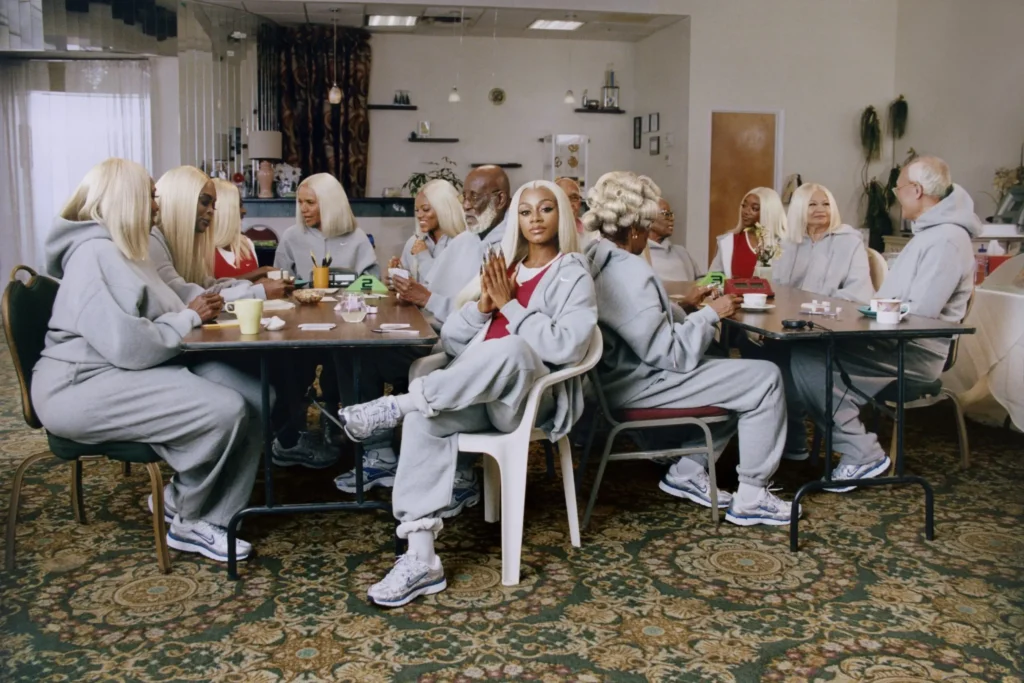 Community center lounge filled with people of different generations wearing coordinated gray Nike fleece, chatting, playing cards, and relaxing around tables while one person sits centered facing the camera