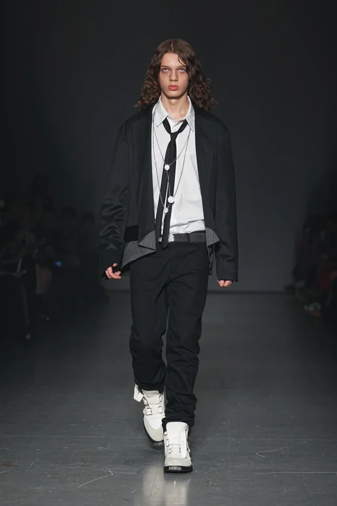 Model wearing a cropped black jacket over a white shirt with a loosened black tie, layered necklaces, slim black trousers, and white buckle sneaker