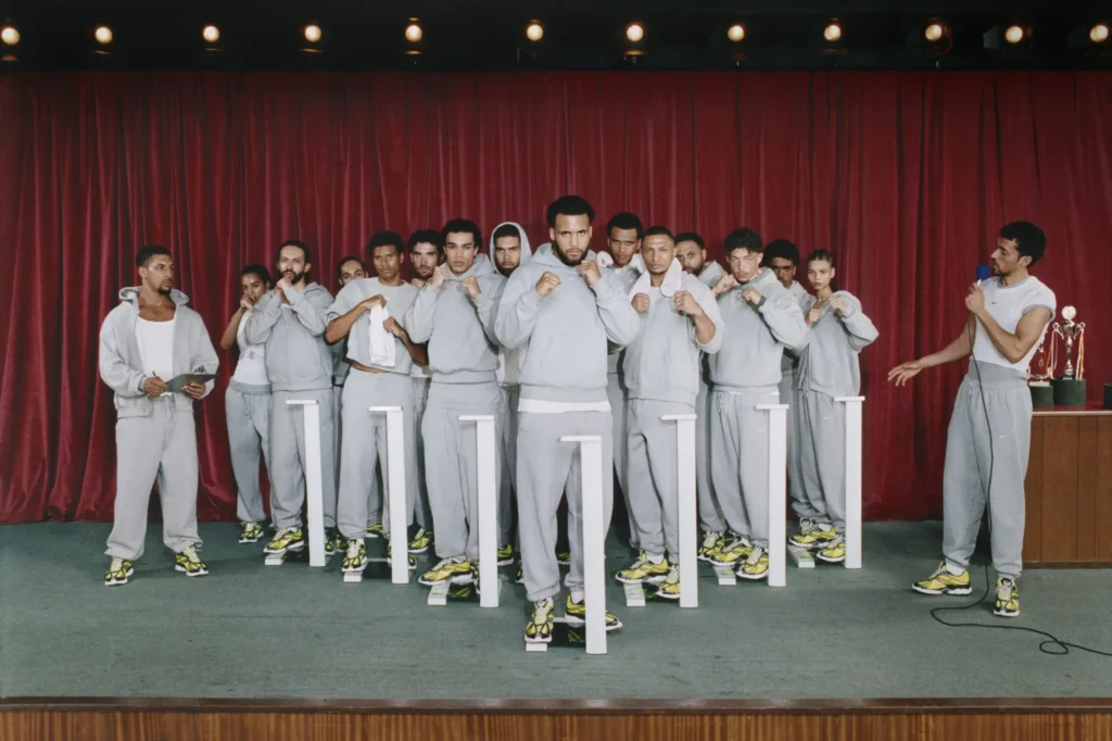 Group of athletes in matching gray Nike fleece tracksuits stand on a stage with boxing stances in front of a red theater curtain, while presenters stand at either side with microphones and clipboard