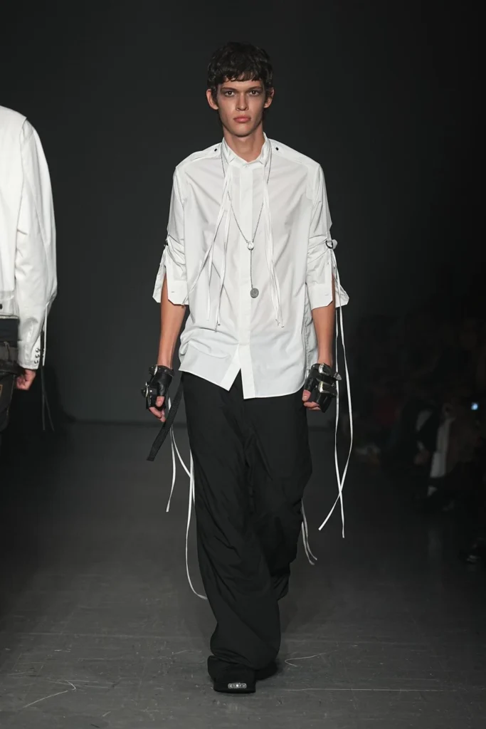 Model in a crisp white shirt with ribbon-tied rolled sleeves, layered pendant necklace, black wide-leg trousers, fingerless gloves, and black shoe