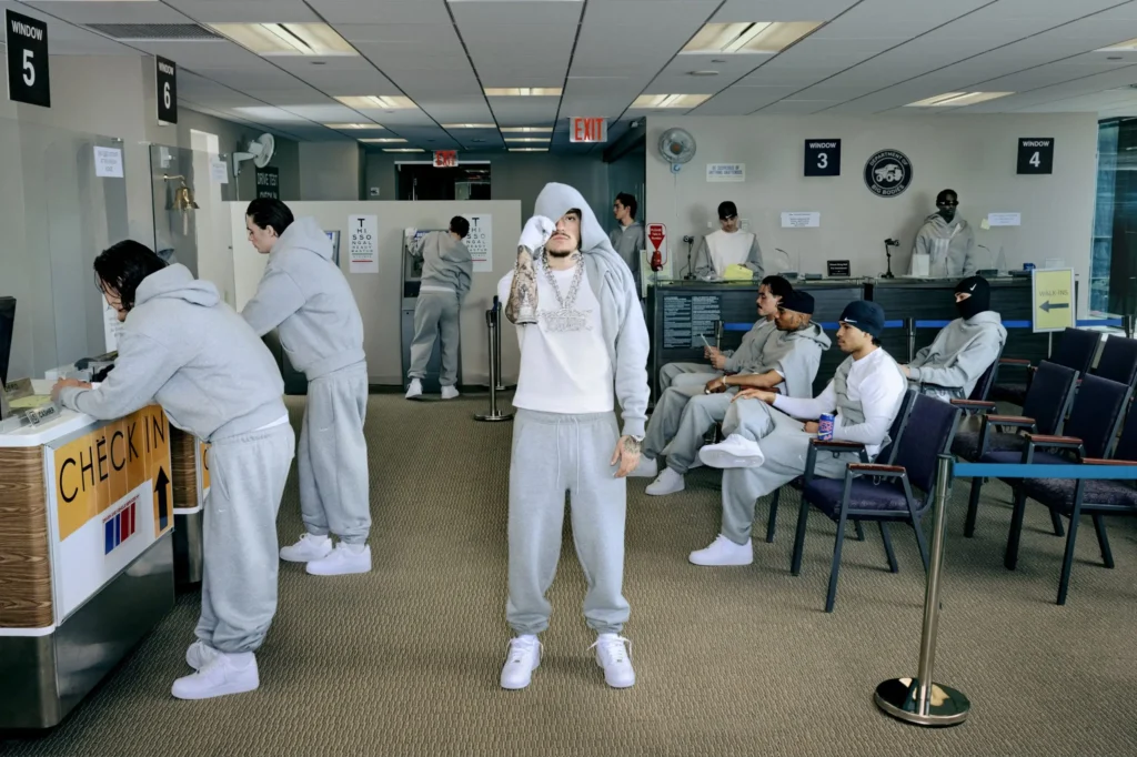 People dressed in coordinated gray Nike fleece wait inside a DMV office, checking in at service counters, sitting in waiting chairs, and standing throughout the lobby while one hooded person poses in the center