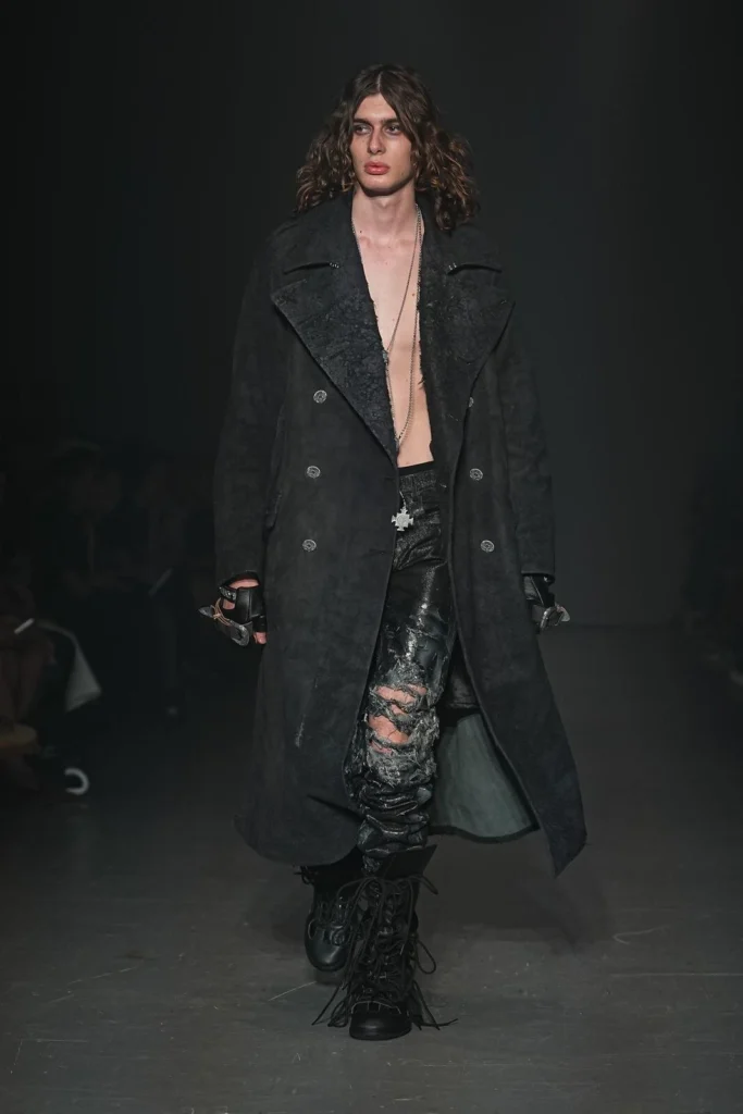 Model wearing a long black double-breasted coat over distressed black trousers, layered necklaces, fingerless gloves, and heavy black lace-up boot
