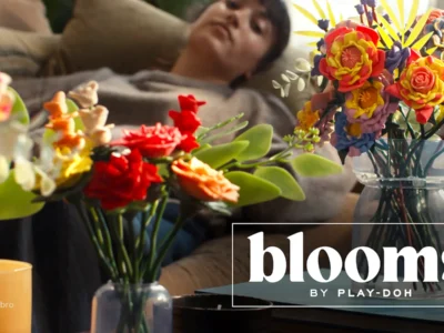 Play-Doh flower bouquets arranged in clear glass vases sit on a coffee table while a person relaxes on a sofa in the background. The image promotes Hasbro's "Blooms by Play-Doh" line, showcasing handcrafted floral designs made entirely from modeling compound in a warm, home set