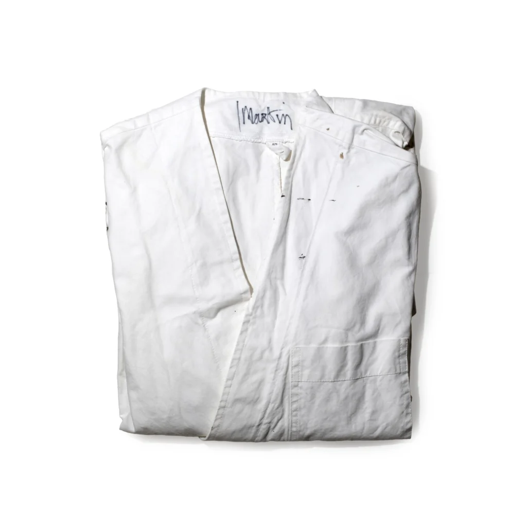 Folded white cotton atelier work coat with subtle paint marks and handwritten "Martin" signature at the collar, photographed as a preserved studio garment