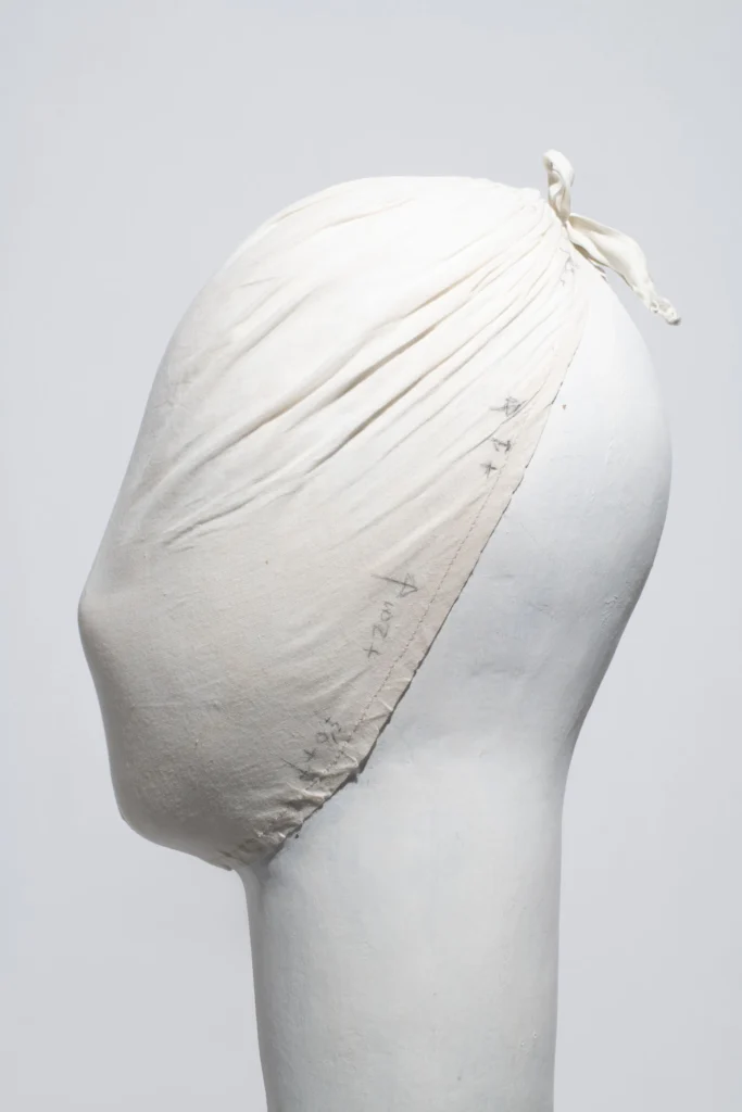White fabric-covered mannequin head wrapped in a tied muslin fitting cap featuring visible pencil markings and tailoring annotation