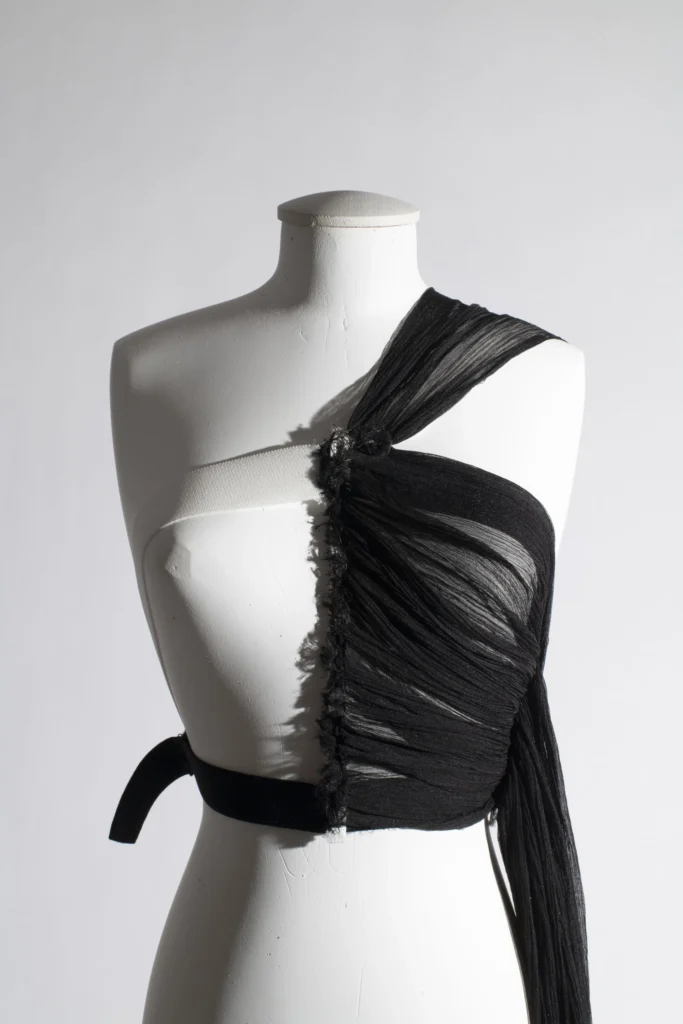 Black asymmetrical draped top made from sheer pleated fabric with exposed white structural strap, displayed on a white mannequin torso
