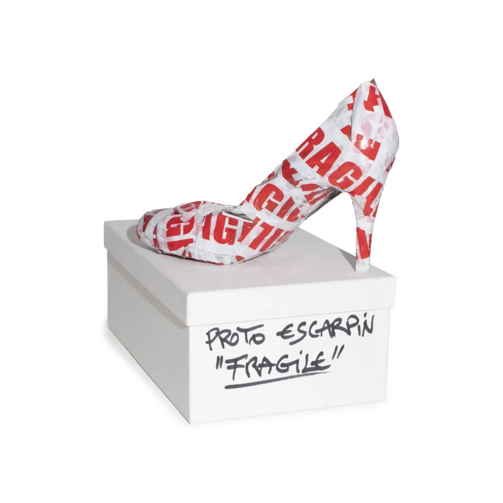 Prototype high-heeled pump wrapped in red-and-white "FRAGILE" tape, displayed atop a labeled white shoebox as an experimental footwear concept