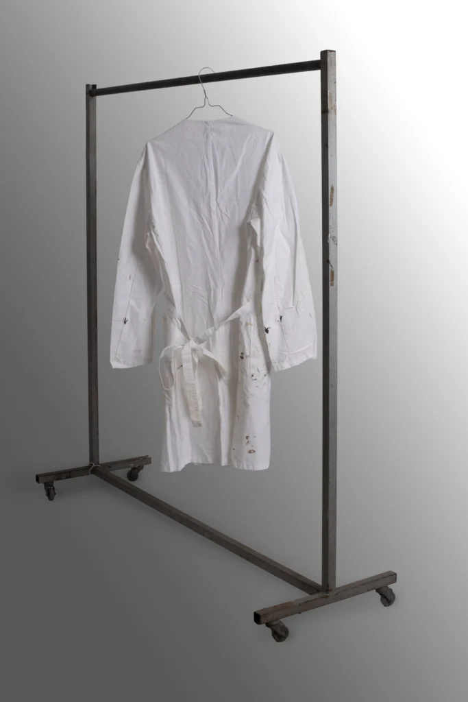 Paint-marked white atelier coat hanging from an industrial rolling garment rack, highlighting the worn workspace aesthetic of a fashion studio