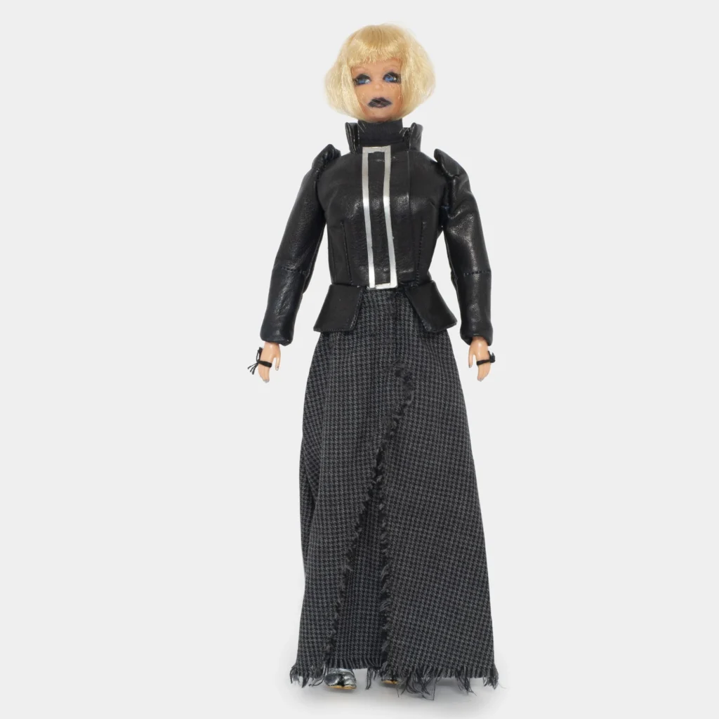 Fashion doll dressed in a structured black jacket with metallic trim, long gray textured skirt, dramatic blue eye makeup, and short blonde bob hairstyle