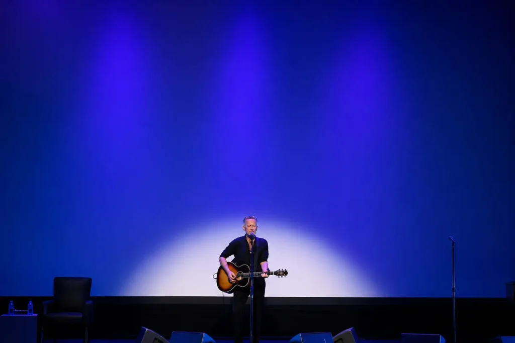 A lone musician stands center stage under deep blue and violet spotlights, singing into a microphone while playing an acoustic guitar. The minimalist stage features a black chair and side table with water bottles on the left, an unused microphone stand on the right, and a large illuminated backdrop that emphasizes the intimate solo show