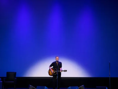 A lone musician stands center stage under deep blue and violet spotlights, singing into a microphone while playing an acoustic guitar. The minimalist stage features a black chair and side table with water bottles on the left, an unused microphone stand on the right, and a large illuminated backdrop that emphasizes the intimate solo show