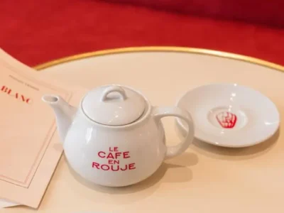 A white ceramic teapot printed with "Le Café en Rouge" sits on a cream café table beside a matching saucer and plate with red branding, while a magazine and red upholstered seating create a warm Parisian café atmosphere