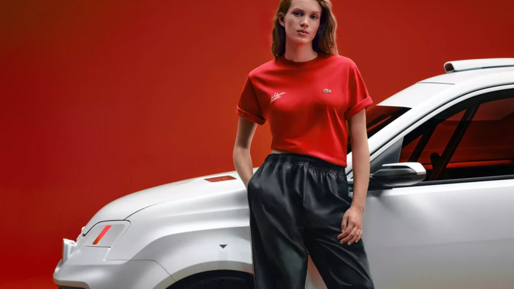 Model wearing a red Alpine x Lacoste T-shirt with embroidered Alpine and Lacoste crocodile logos, standing beside the silver Alpine A290 Rallye against a vivid red studio backdrop