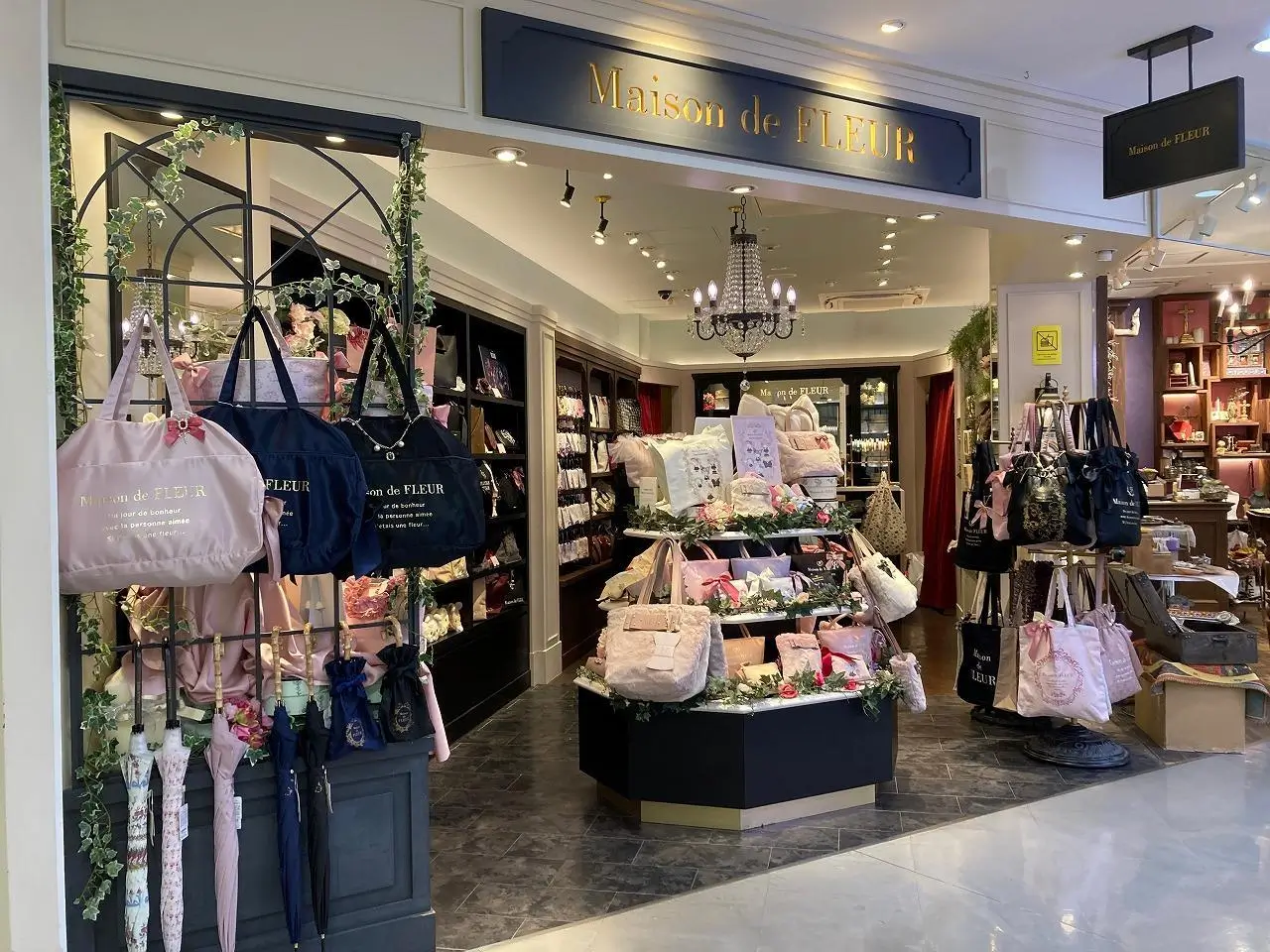 Maison de FLEUR storefront featuring an elegant boutique entrance with pastel tote bags, floral displays, plush accessories, umbrellas, and home décor arranged beneath black-and-gold signage and chandelier lighting.