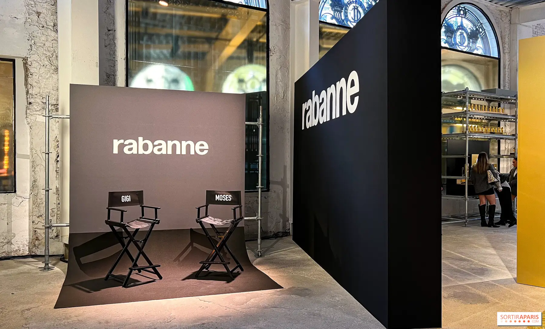 Interior of a Rabanne-branded pop-up installation featuring oversized black and charcoal display walls with bold white logos, two black director's chairs labeled 'Gigi' and 'Moses' on a small stage, and an industrial venue framed by tall arched windows and exposed concrete finishes.