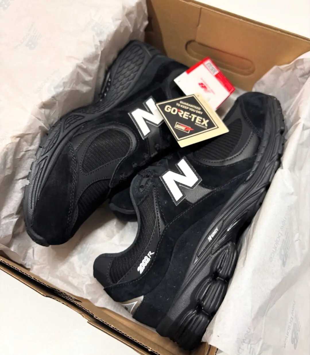 New Balance 2002R GORE-TEX sneakers in an all-black colorway resting inside an open shoebox with tissue paper, showcasing black suede and mesh construction, white 'N' logos, ABZORB cushioning, and a hanging GORE-TEX waterproof tag.