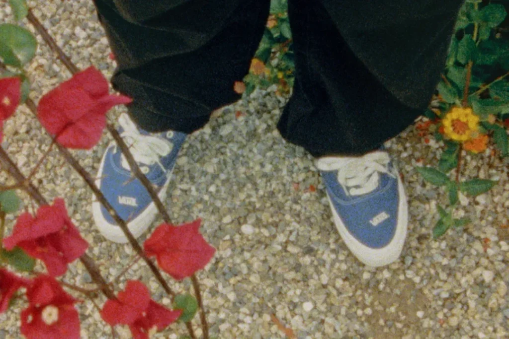 Person wearing blue Vans OTW low-top canvas sneakers with white laces and white foxing, standing on a pebled garden path surrounded by red bougainvillea and yellow flowers in a softly grainy, vintage-style photograph