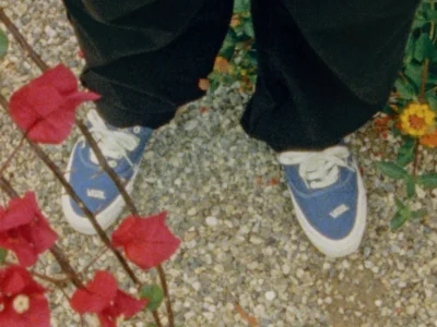 Person wearing blue Vans OTW low-top canvas sneakers with white laces and white foxing, standing on a pebled garden path surrounded by red bougainvillea and yellow flowers in a softly grainy, vintage-style photograph