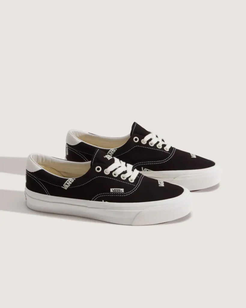 Pair of black Vans OTW low-top canvas shoe with scattered white Vans logos, contrast white stitching, white laces, and thick white rubber soles displayed on a light neutral studio background