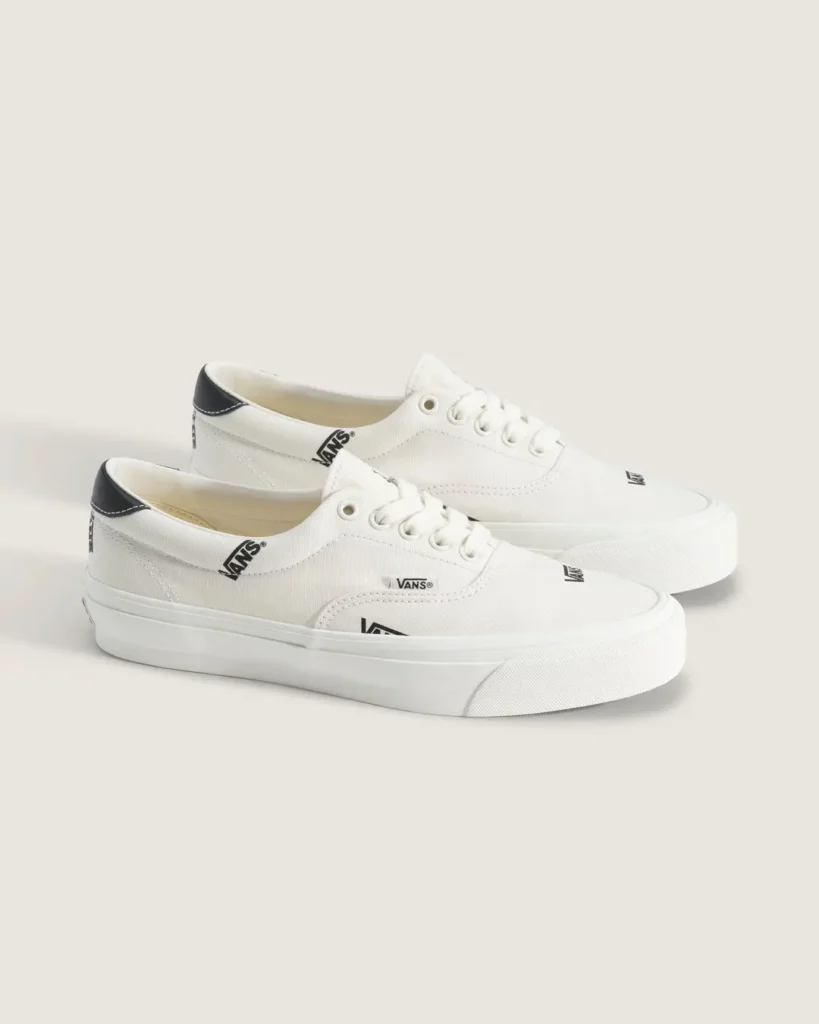 Pair of white Vans OTW low-top canvas shoe with black Vans logo prints, black leather heel tabs, white laces, and white vulcanized soles shown against a minimalist beige backdrop