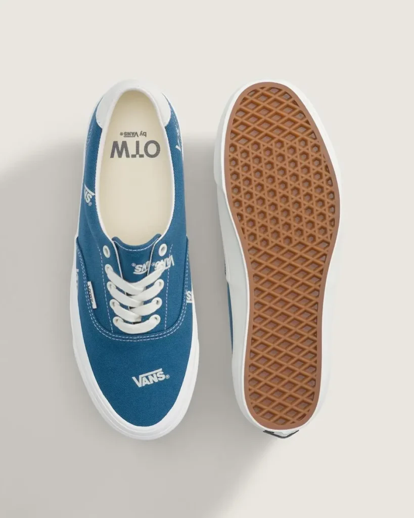 Pair of white Vans OTW low-top canvas sneakers with black Vans logo prints, black leather heel tabs, white laces, and white vulcanized soles shown against a minimalist beige backdrop